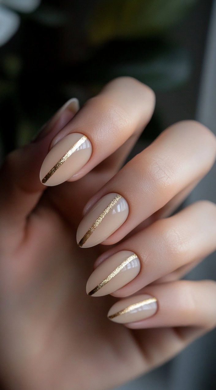 A close-up of a hand showcasing almond-shaped nude nails with a sleek gold stripe running down the center of each nail.