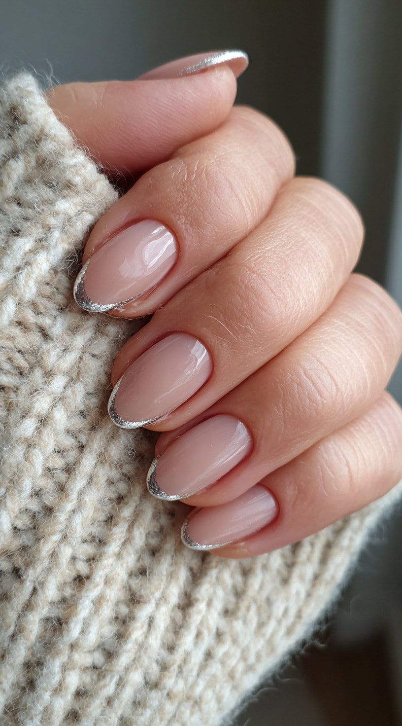 Almond-shaped nude nails with subtle silver glitter French tips over a sheer base.