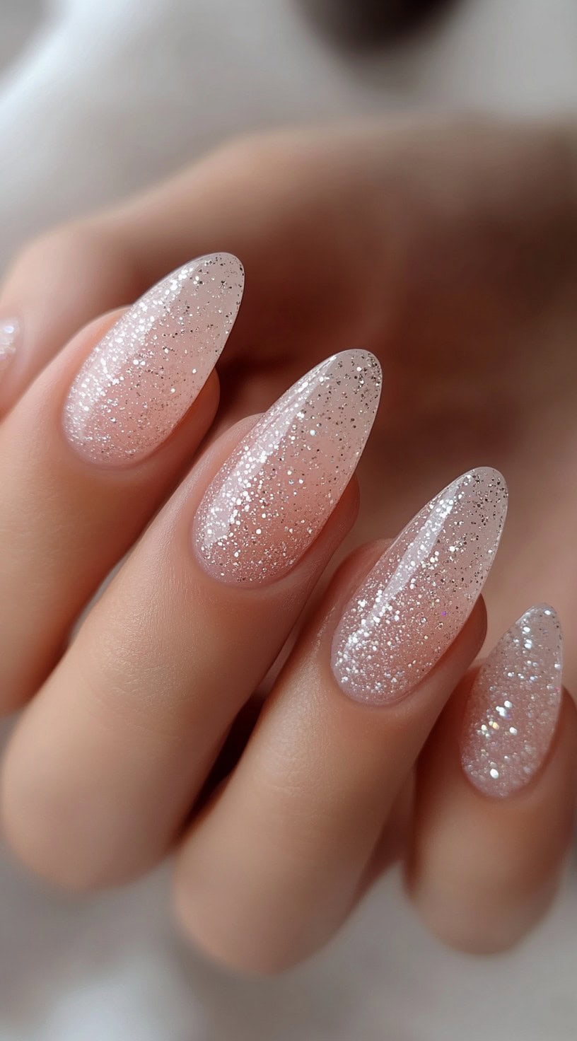 A set of almond-shaped nails with a soft nude-to-silver ombre glitter effect.