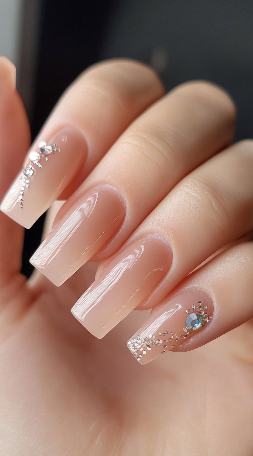 A hand with long, square-shaped nails in a soft nude-pink shade, embellished with small crystal accents.