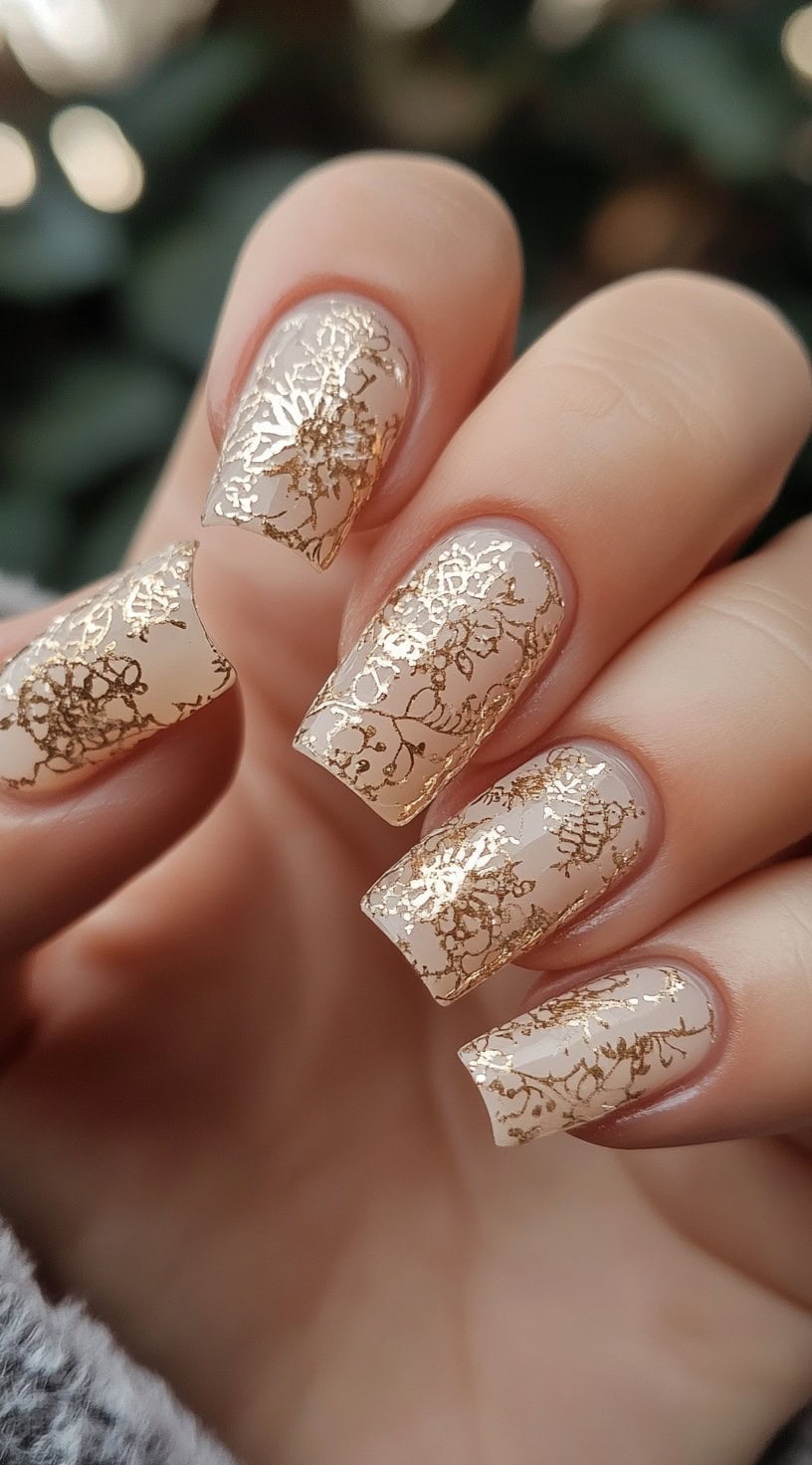 A hand with square-shaped nude nails featuring intricate gold lace patterns for an elegant, vintage-inspired look.