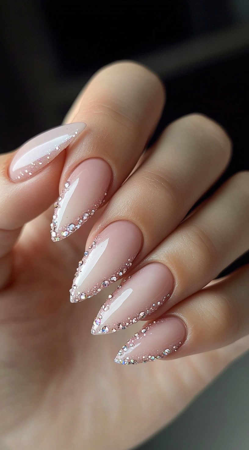 A close-up of stiletto-shaped nails with a nude base, adorned with a delicate row of sparkling crystals along the French tips.