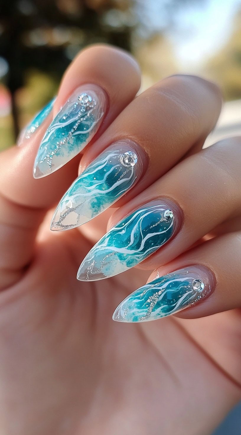 Stiletto-shaped nails with a marbled blue and white wave pattern and silver gem accents.