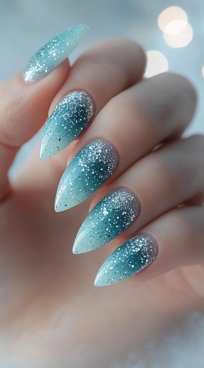 Almond-shaped nails with a teal blue ombre base fading into silver glitter near the cuticles.