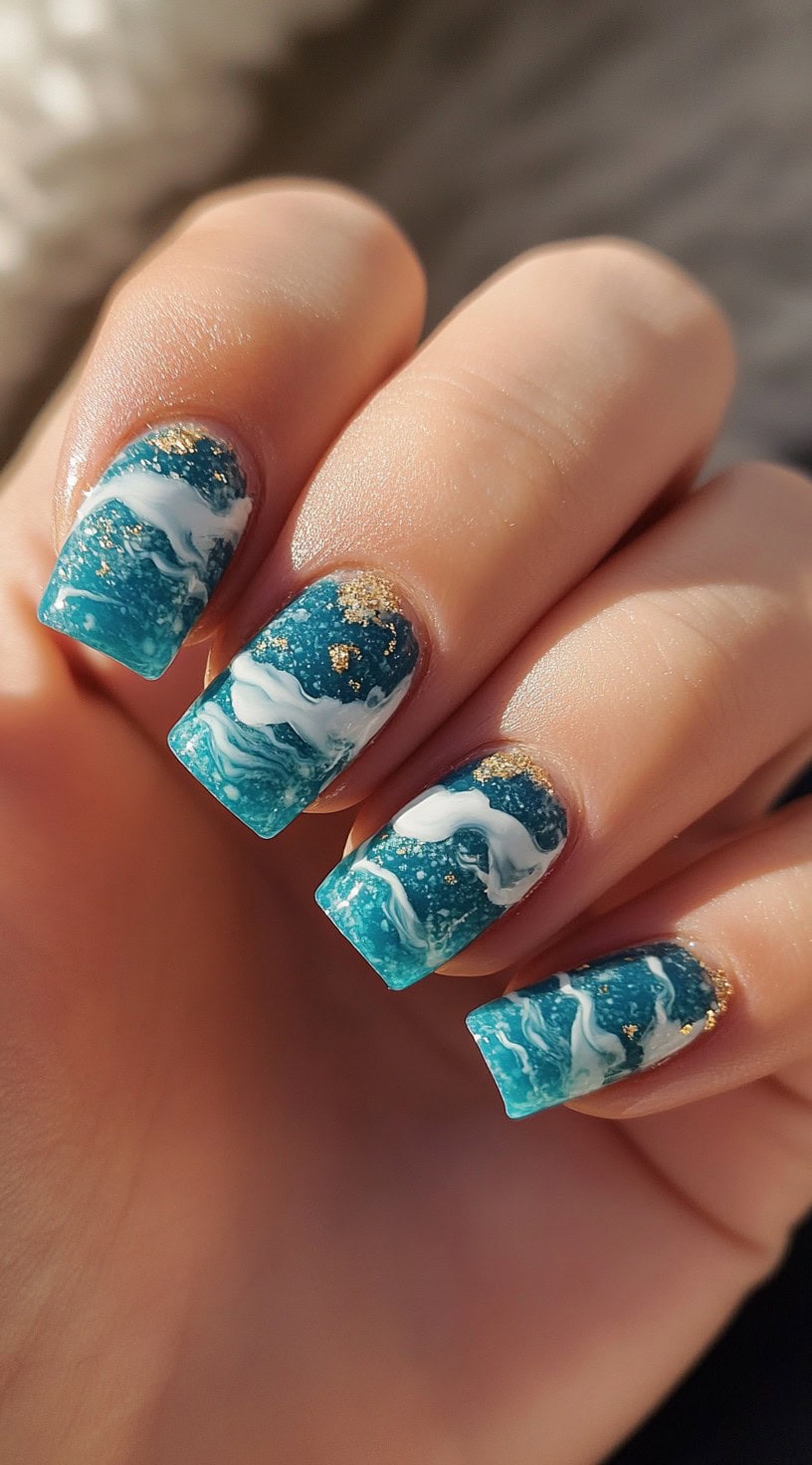 Square nails painted in ocean blue with white wave accents and scattered gold flakes.