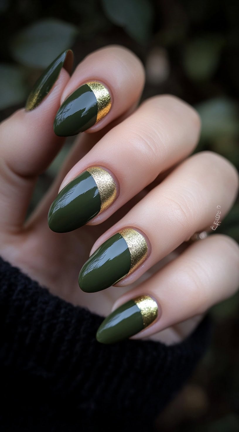 A hand with almond-shaped nails featuring a deep olive green polish and a bold gold half-moon design at the base, creating a luxurious contrast.