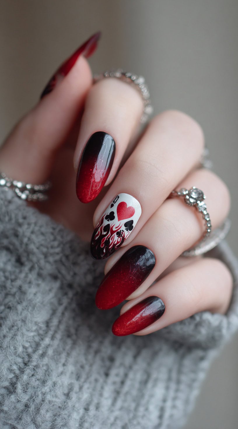 A hand with long coffin nails featuring black-to-red ombré and one white accent nail with card suits, a red heart, and fiery flames.