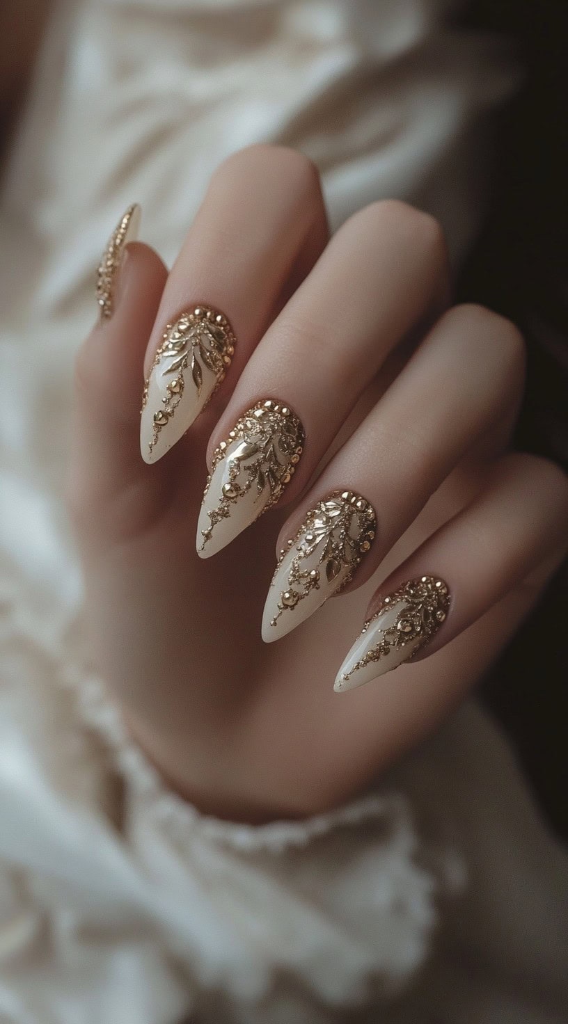 A set of long, almond-shaped nails with an ivory base, adorned with intricate gold beadwork and baroque-style embellishments.