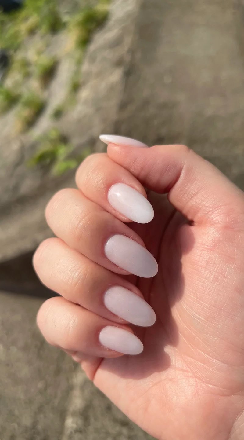 Close-up of short fingers with oval-shaped nails painted in a clean white ombre finish, glowing in natural sunlight.