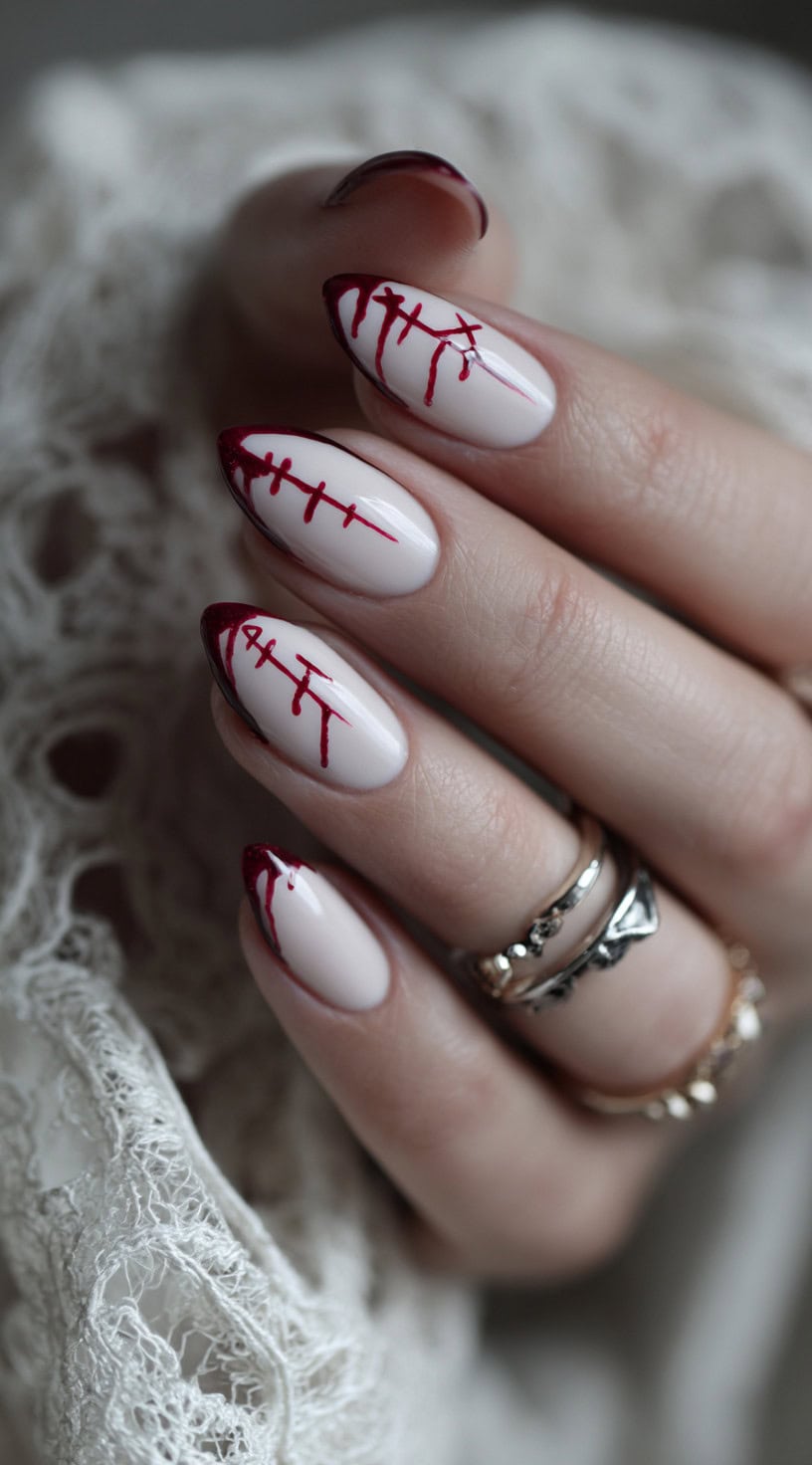 A hand with almond-shaped pale pink nails featuring blood-red rune designs dripping from the French tips.