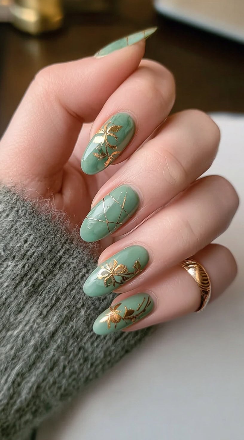 A hand with soft almond-shaped nails in pale jade, decorated with delicate gold foil flowers and line detailing.