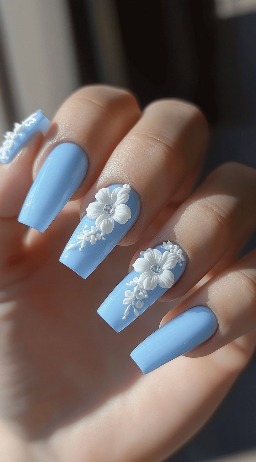 Pastel blue coffin nails with intricate 3D white floral designs and rhinestone embellishments.