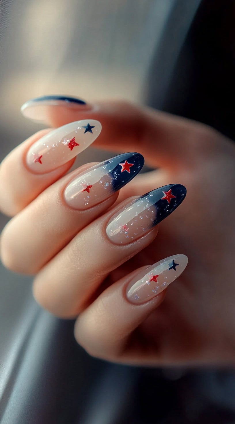 A hand with long almond-shaped nails featuring sheer pink bases, deep blue French tips, and red and blue star accents with subtle glitter.