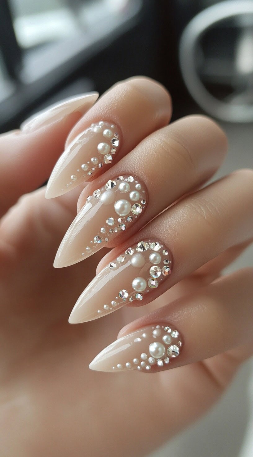A close-up of long almond-shaped nails with a glossy nude base, decorated with pearls and crystals in a cascading pattern.