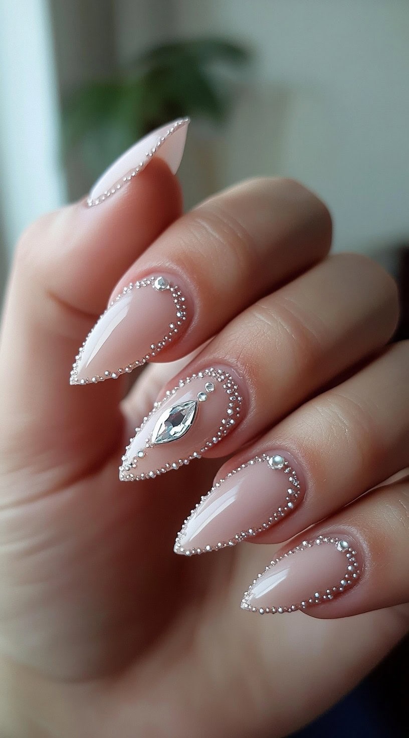 Hand with long almond nails in soft nude, each outlined with tiny pearls and one nail featuring a large crystal gem.