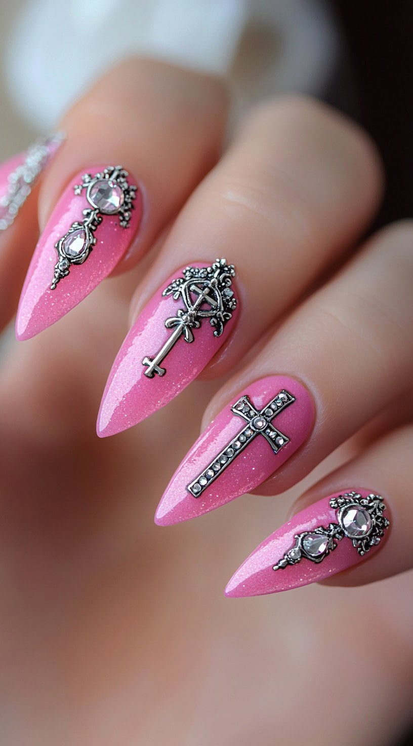 A hand with long pink almond nails adorned with ornate silver cross and crystal charms.