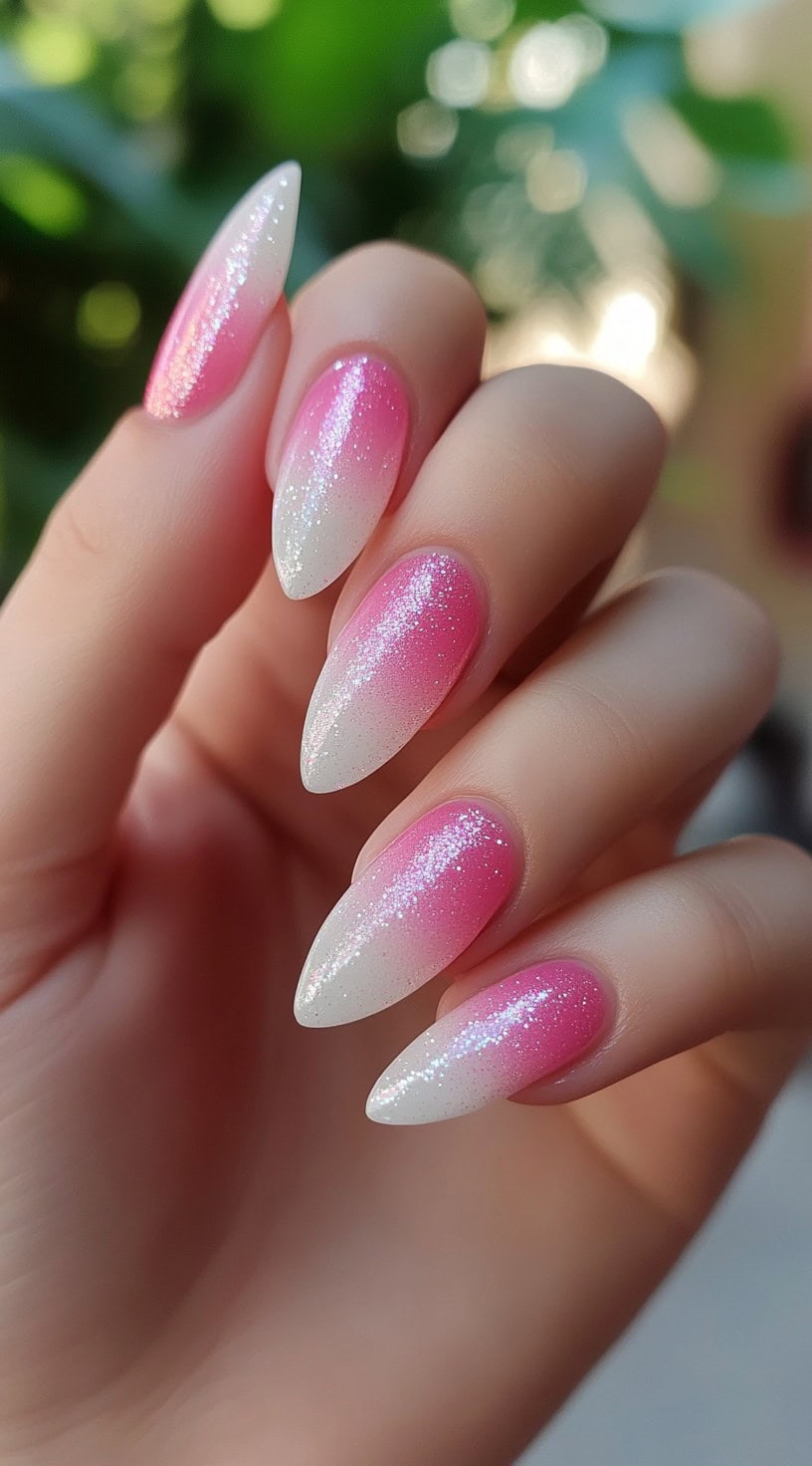 A hand with almond-shaped nails featuring a pink-to-white gradient infused with ultra-fine glitter, creating a soft and luminous effect.