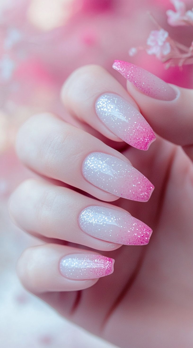 A hand with long square nails showing a smooth transition from soft white to bright pink, topped with sparkling glitter and high-gloss shine.