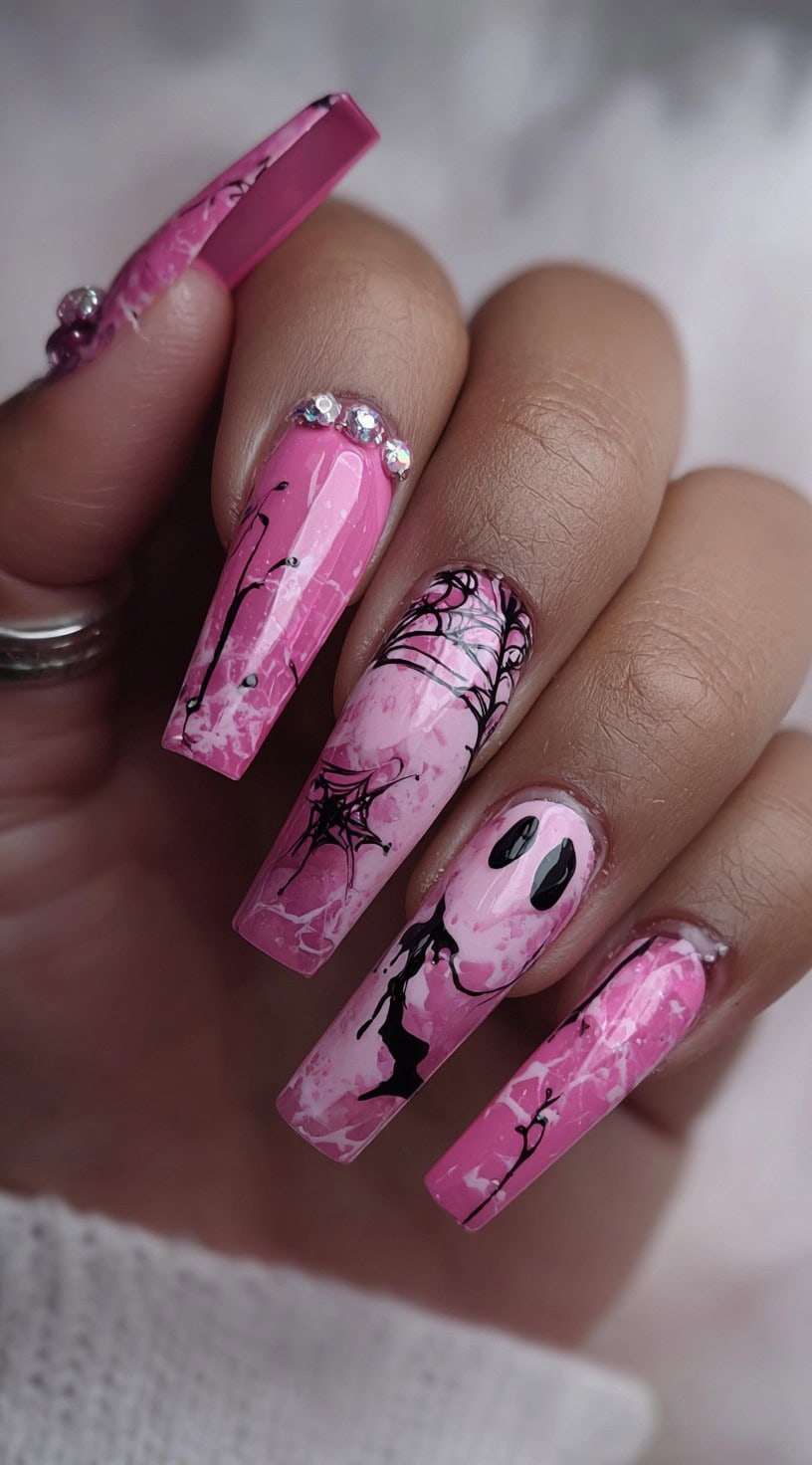 Hand with long pink coffin nails featuring marbled patterns, black spiderwebs, a ghost face, and rhinestone embellishments.