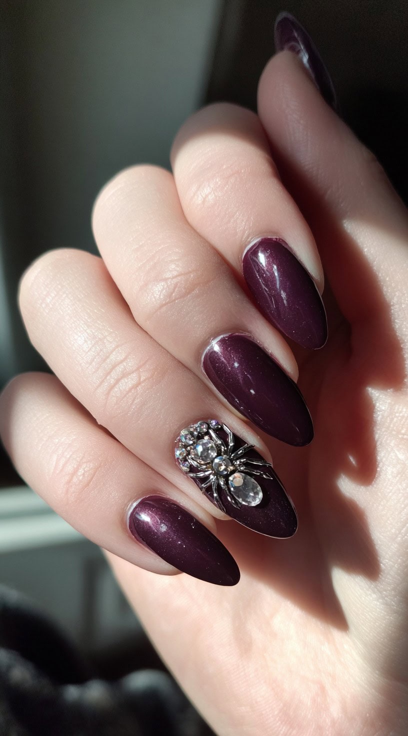 A hand with glossy plum-colored nails, featuring a sparkling rhinestone spider on the ring finger.
