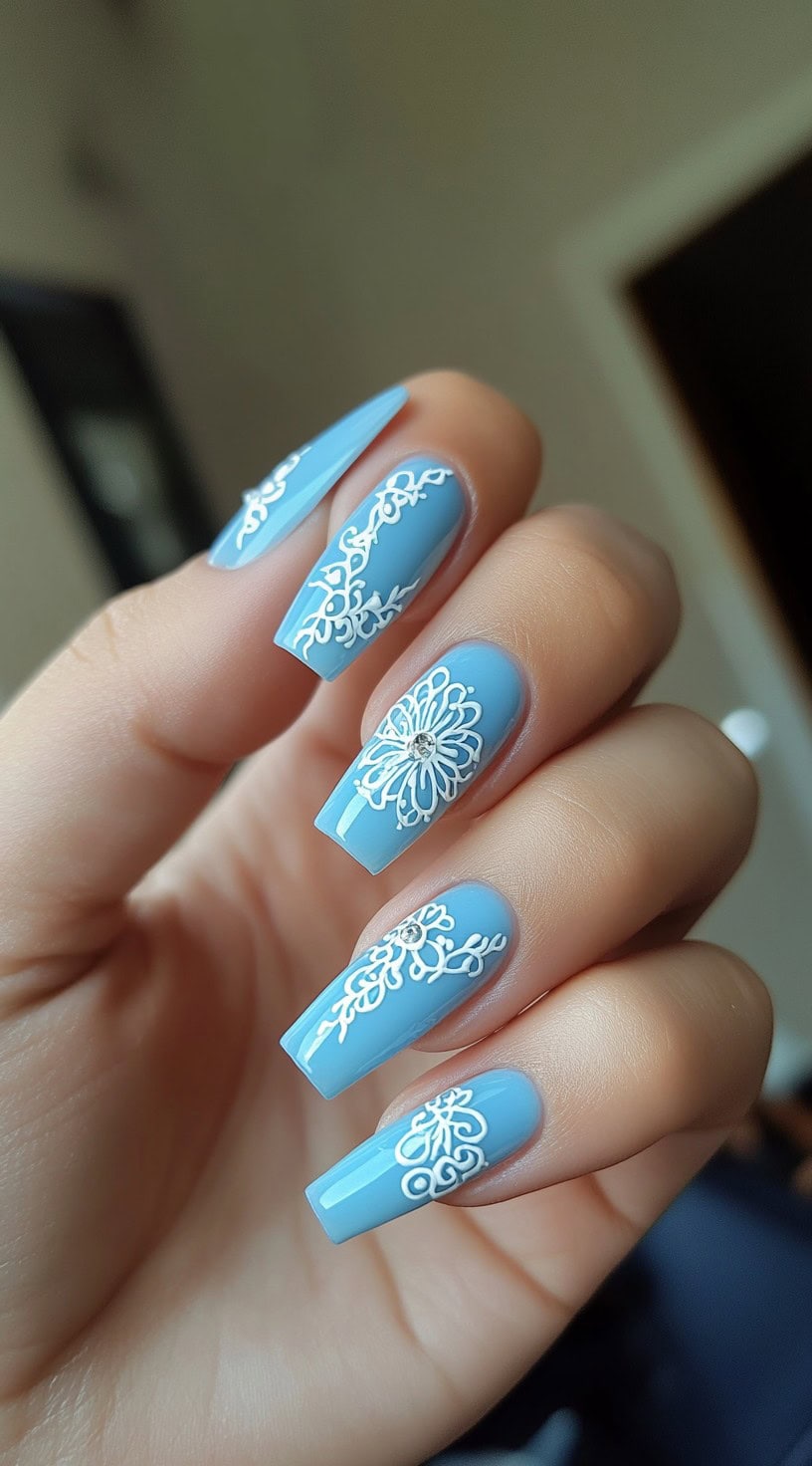Five powder blue coffin-shaped nails with ornate white lace detailing and small rhinestone embellishments at the center of some designs.