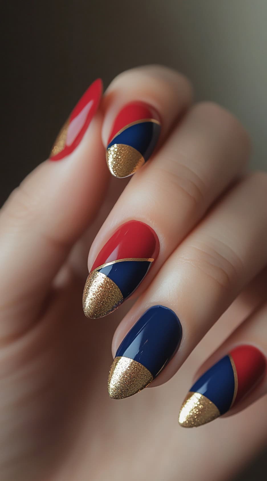 A hand displaying almond-shaped nails with a red and blue French tip design, separated by elegant gold detailing, with gold-glittered tips for extra glamour.