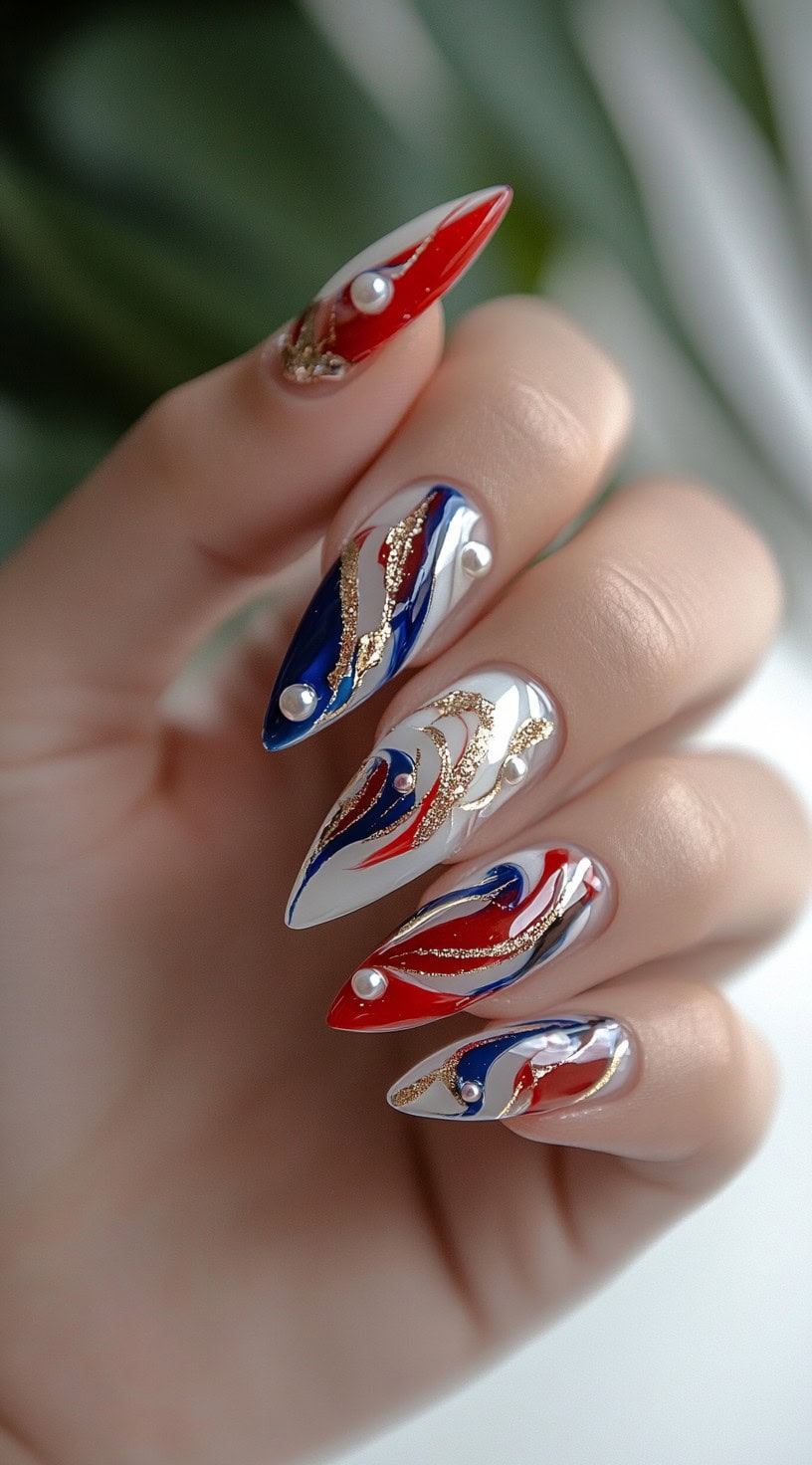 A hand showcasing long, stiletto nails with an intricate red, blue, and gold swirl design, accented with pearl embellishments and gold foil details.