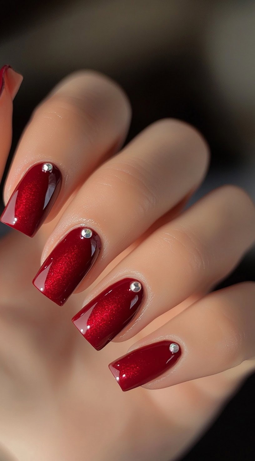 Shimmering deep red square nails with a glossy finish and single rhinestones at the base of each nail.