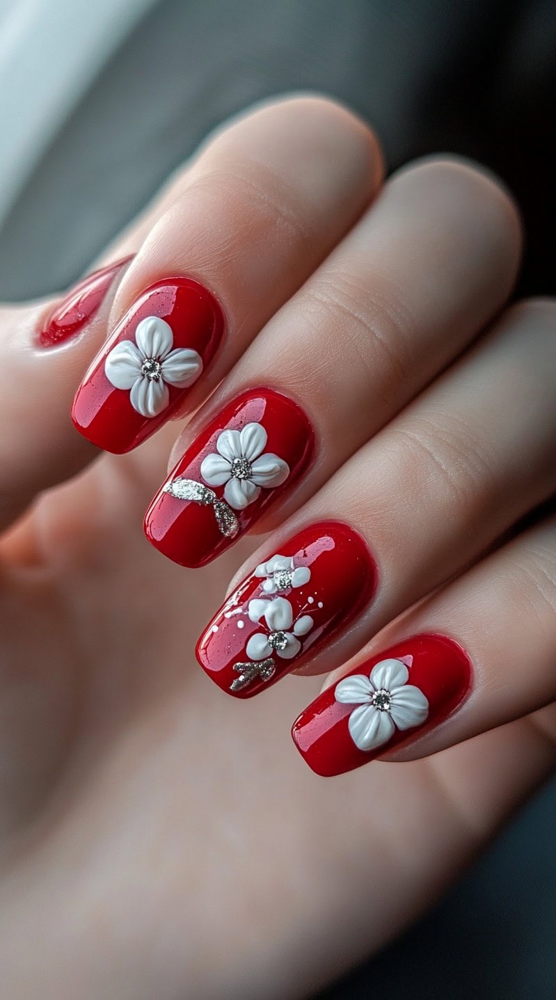 A set of glossy red nails with delicate 3D white floral accents and rhinestone embellishments on a square nail shape.