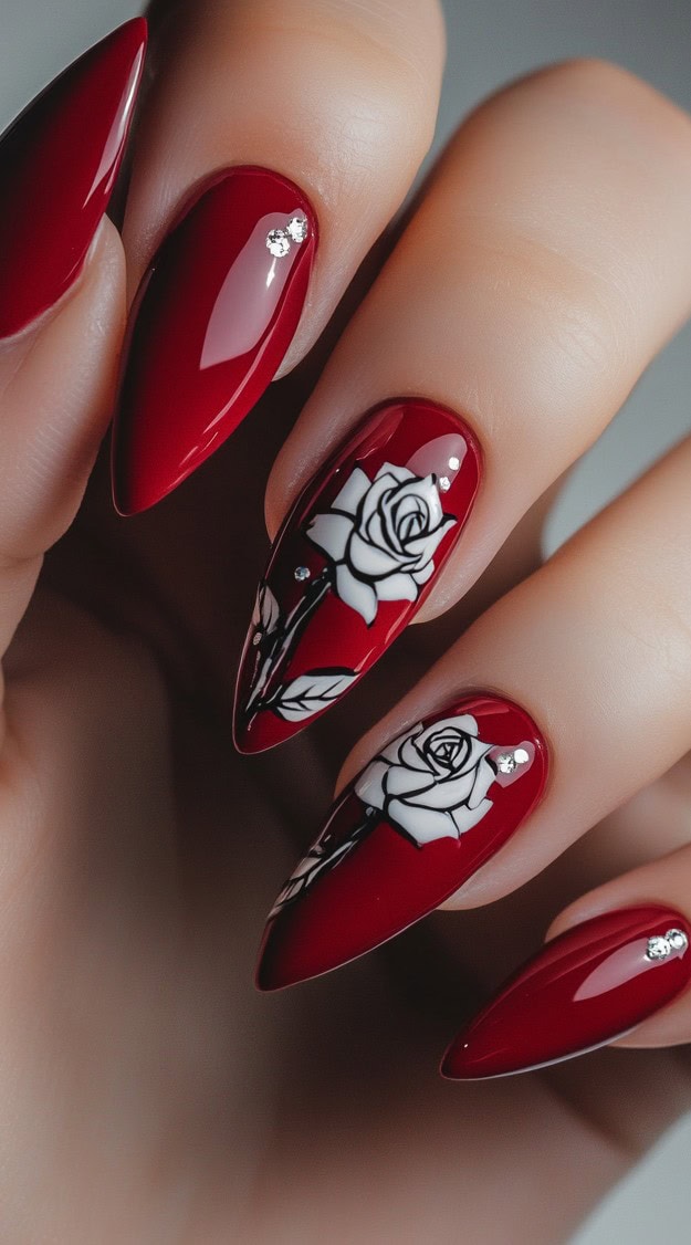 A bold red stiletto manicure with glossy shine, featuring delicate hand-painted white roses and rhinestone embellishments.