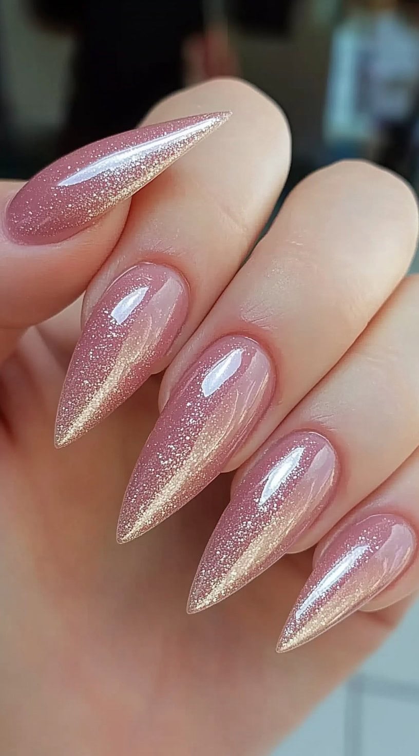 A hand with long stiletto-shaped nails featuring a soft pink base with a rose gold glitter gradient towards the tips, finished with a glossy top coat.