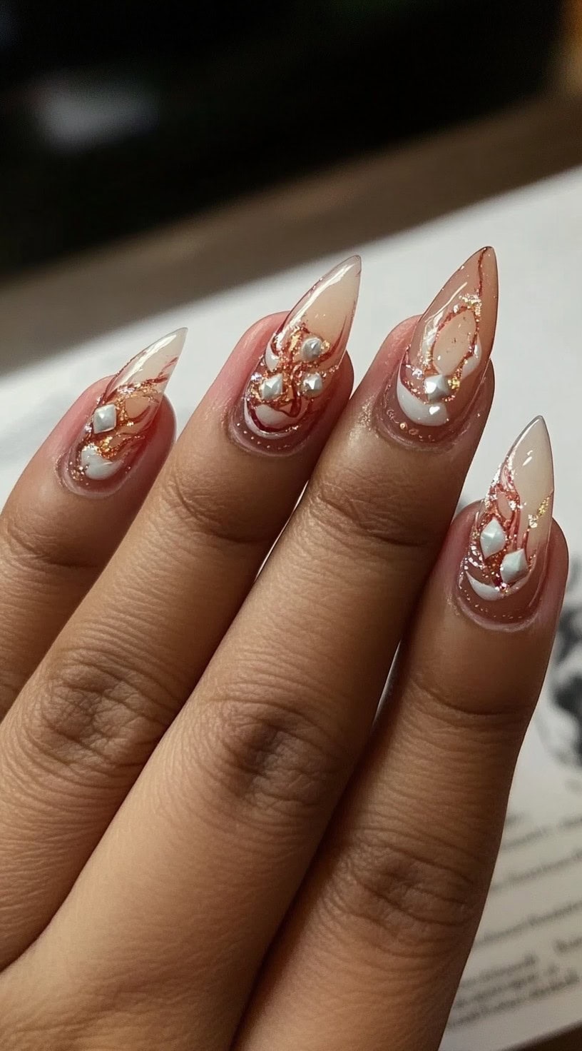 Hand with sharp stiletto nails in nude polish, layered with rose gold foil, white swirls, and raised pearl-like gems.