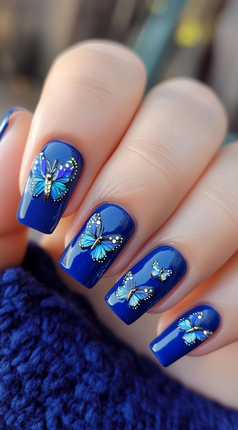 A hand adorned with deep royal blue nails featuring intricate 3D butterfly decals in shades of blue and gold.