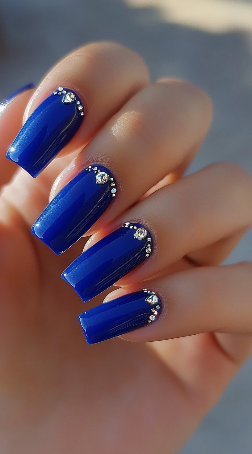 Glossy royal blue coffin nails adorned with sparkling rhinestones near the cuticles.