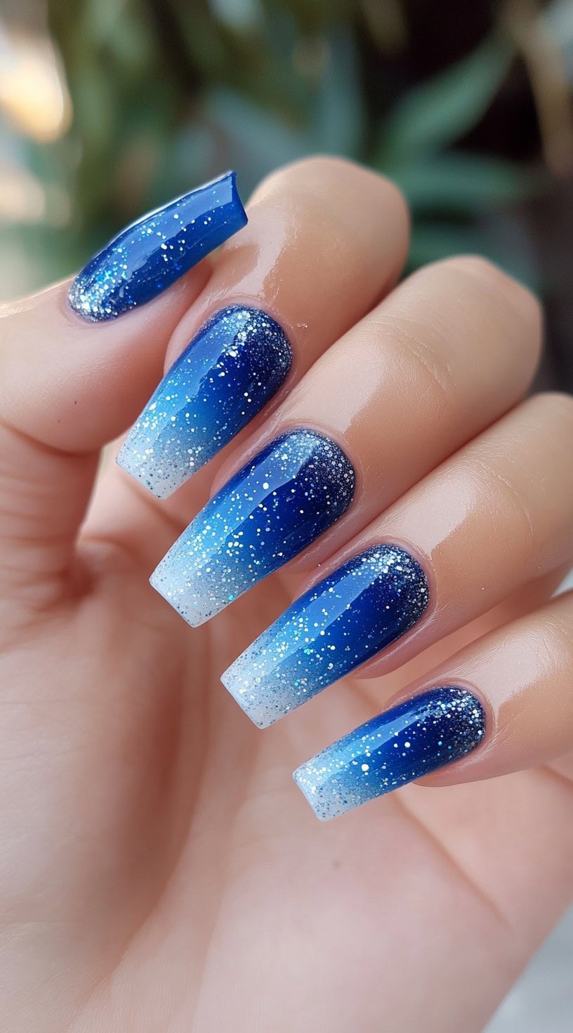 Long coffin-shaped nails with a deep royal blue to icy white ombre and scattered silver glitter.