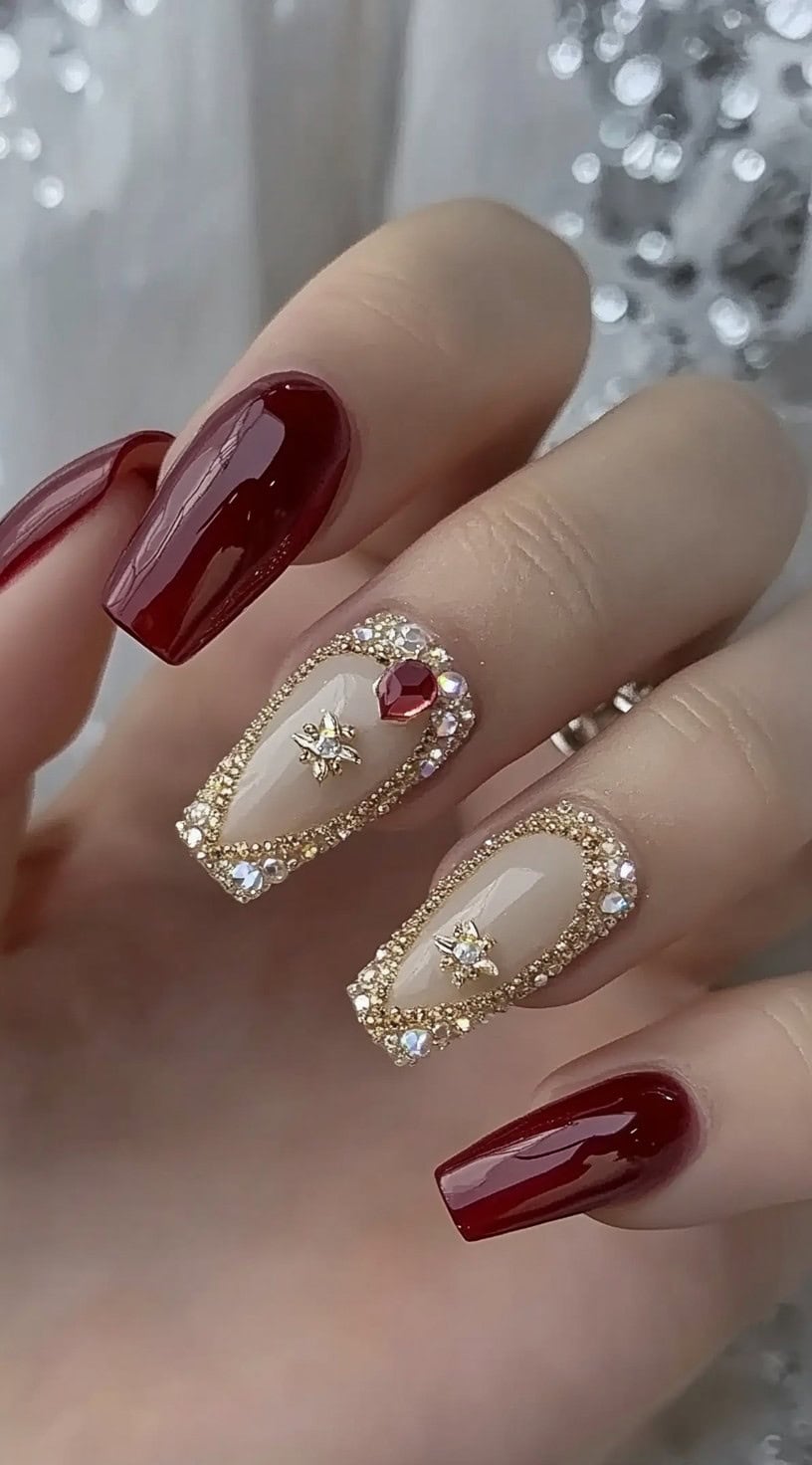 A regal set of burgundy and nude nails adorned with intricate gold embellishments, rhinestones, and a ruby-red accent gem.