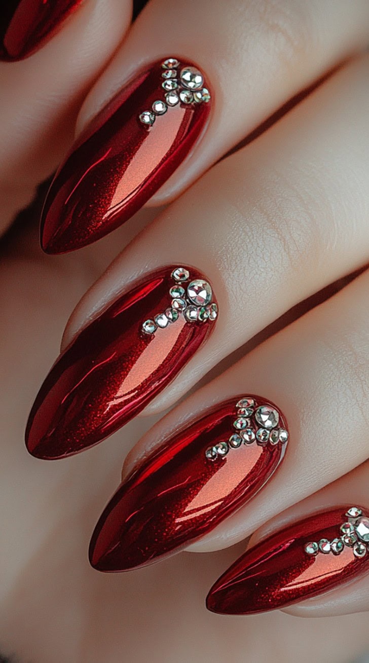 A set of almond-shaped nails painted in a vibrant ruby red, with small silver rhinestones forming elegant patterns along the cuticle and nail bed.