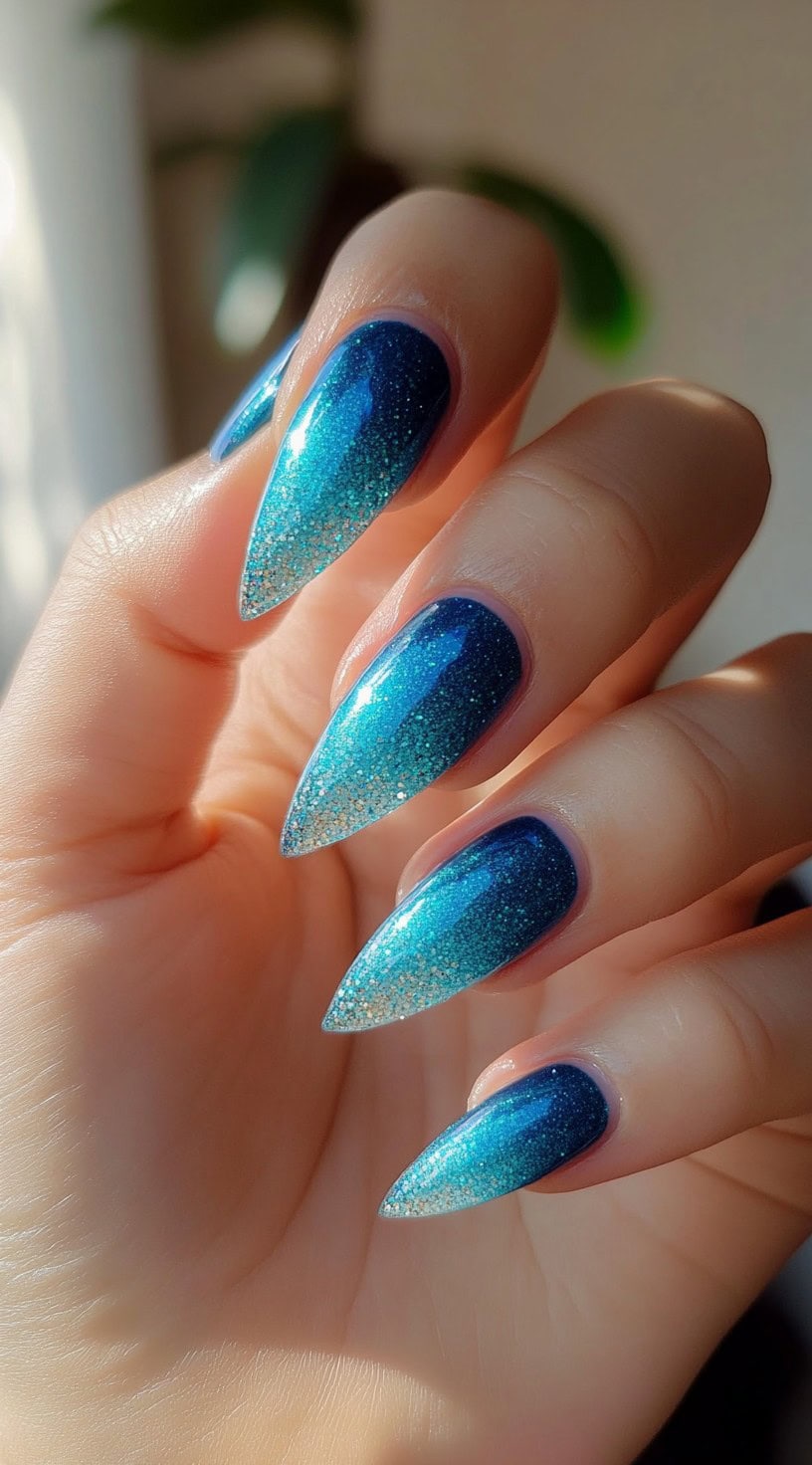 A hand with long stiletto-shaped nails featuring a deep blue to turquoise glitter ombre fade.