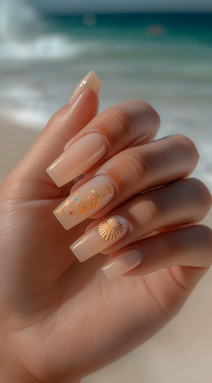 Long square-shaped nails with a light nude polish, adorned with golden sparkles and a single seashell accent nail, with the ocean in the background.