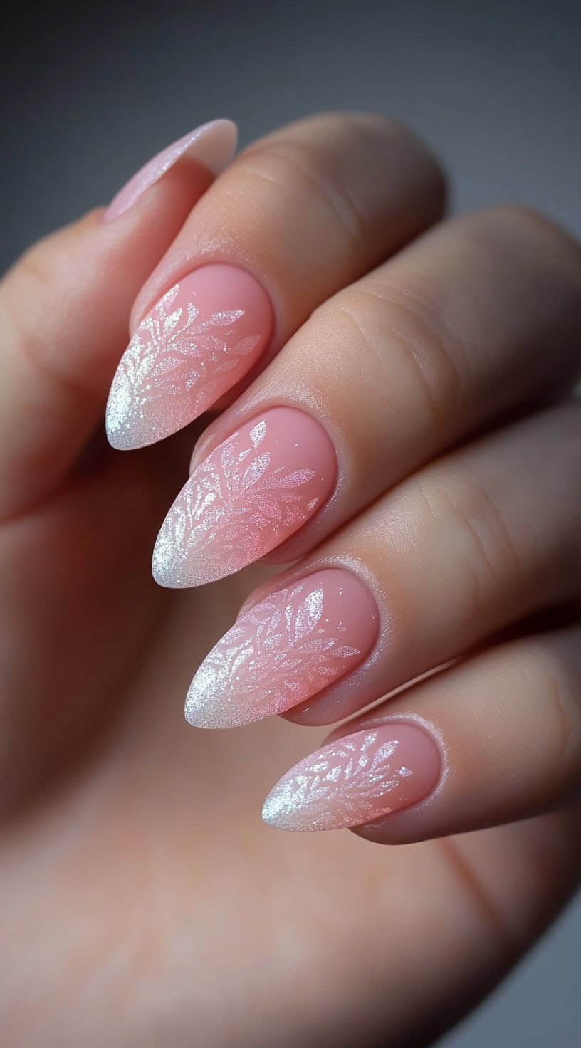 A hand with almond nails in a soft pink ombré, finished with shimmery leaf designs in white.