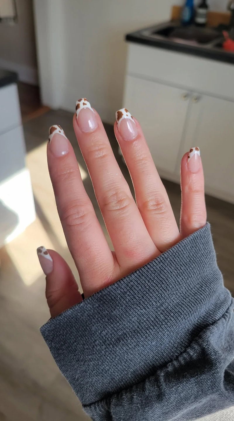Short coffin-shaped nails with clear bases and cow-print French tips, worn on a hand with short fingers and cozy sleeve styling.