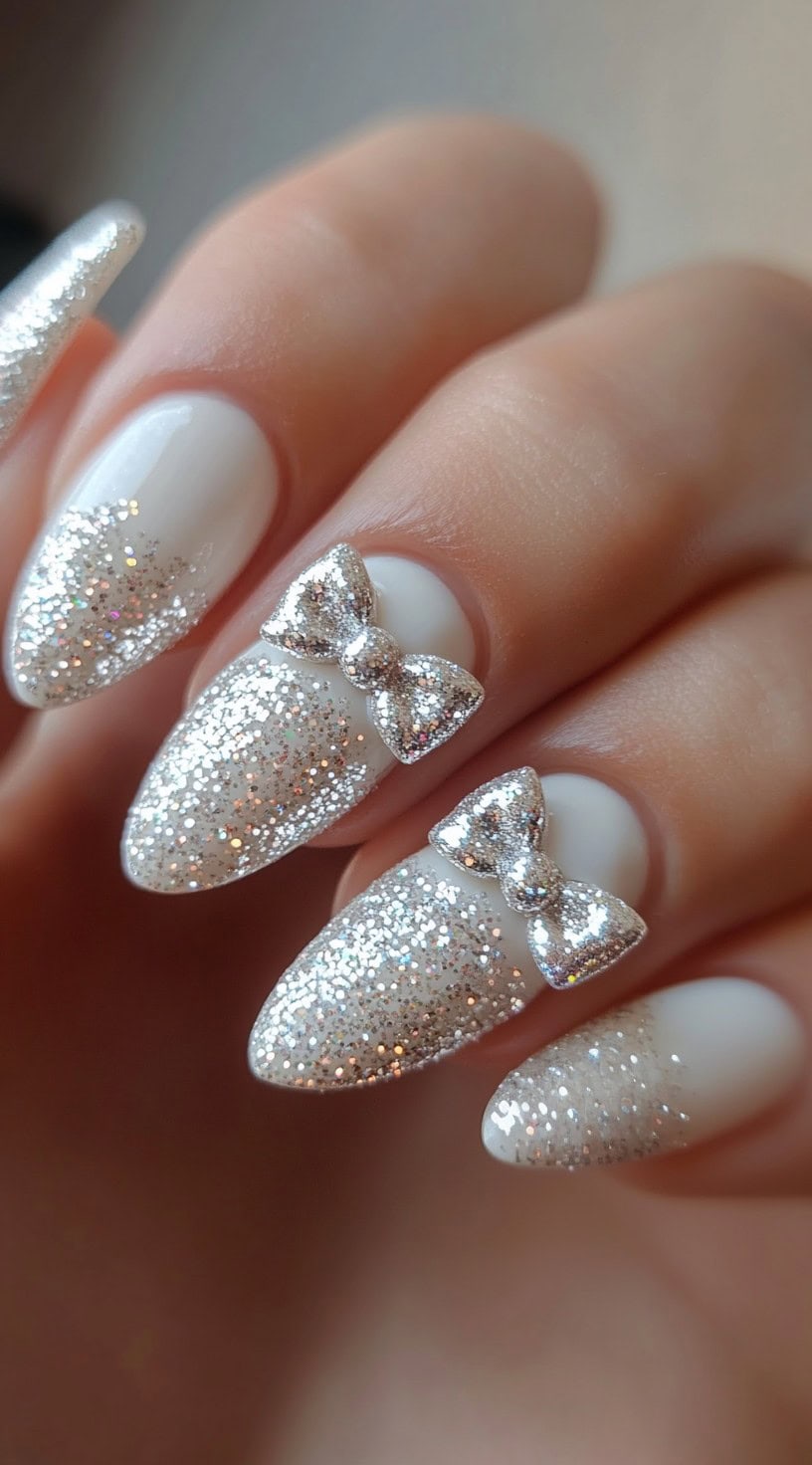 A hand with almond-shaped nails featuring a white base, sparkling silver glitter tips, and 3D silver bow embellishments for a charming, bridal-inspired look.