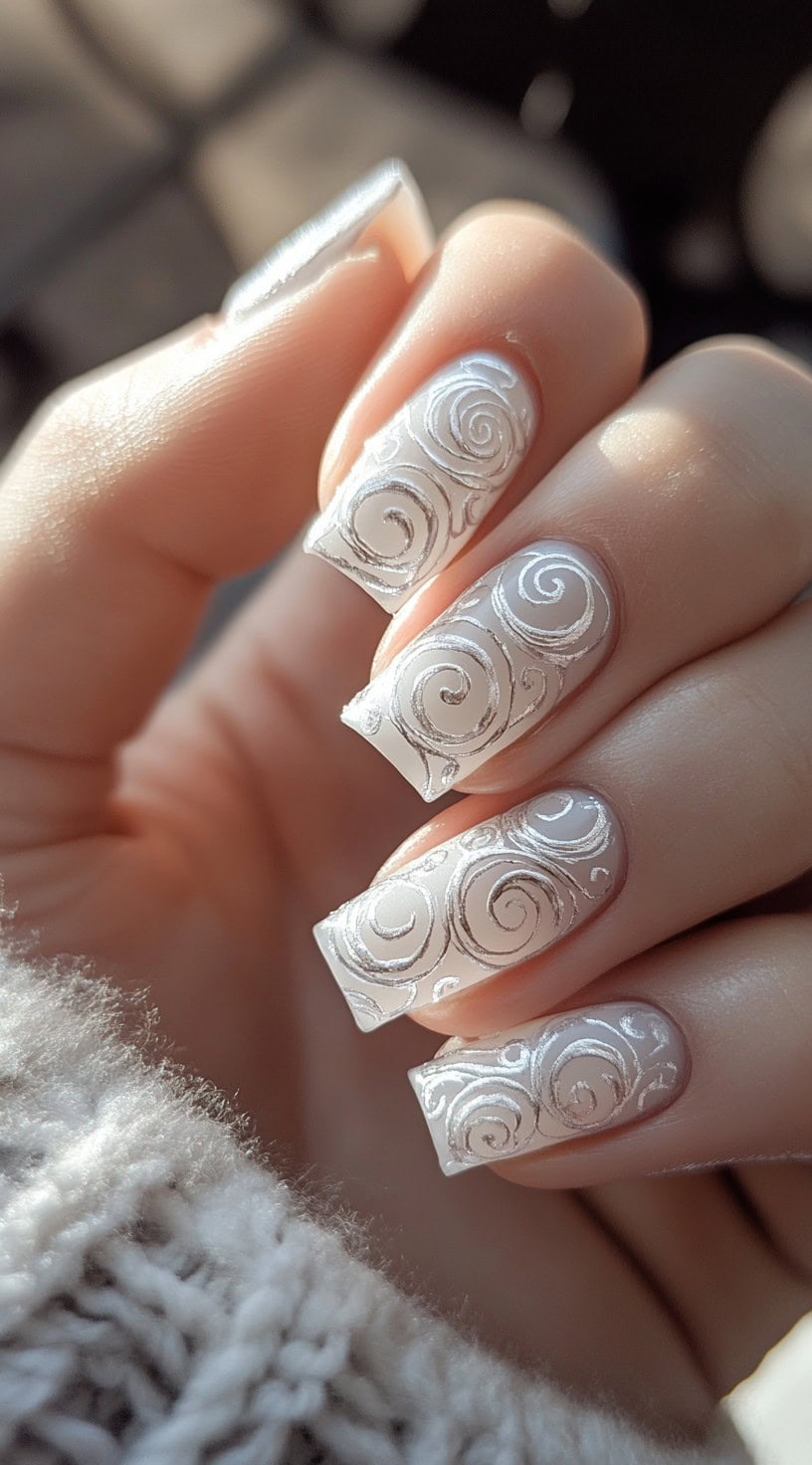 A close-up of square-shaped nails with a white matte base, embossed with delicate silver swirl patterns.