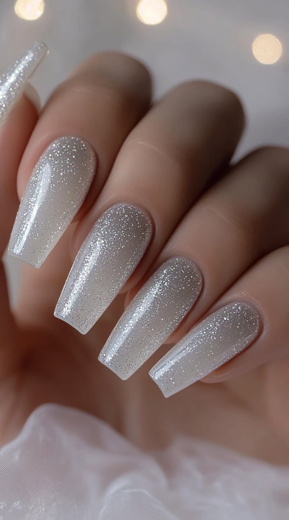 A close-up of long coffin-shaped nails with a sheer nude base transitioning into fine silver glitter towards the tips.