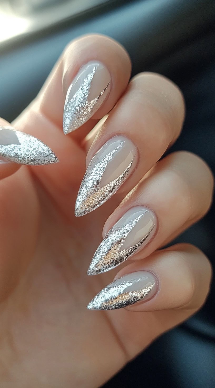 A hand with long, stiletto-shaped nails featuring a sleek, nude base with bold silver metallic glitter streaks forming a dynamic, edgy pattern.