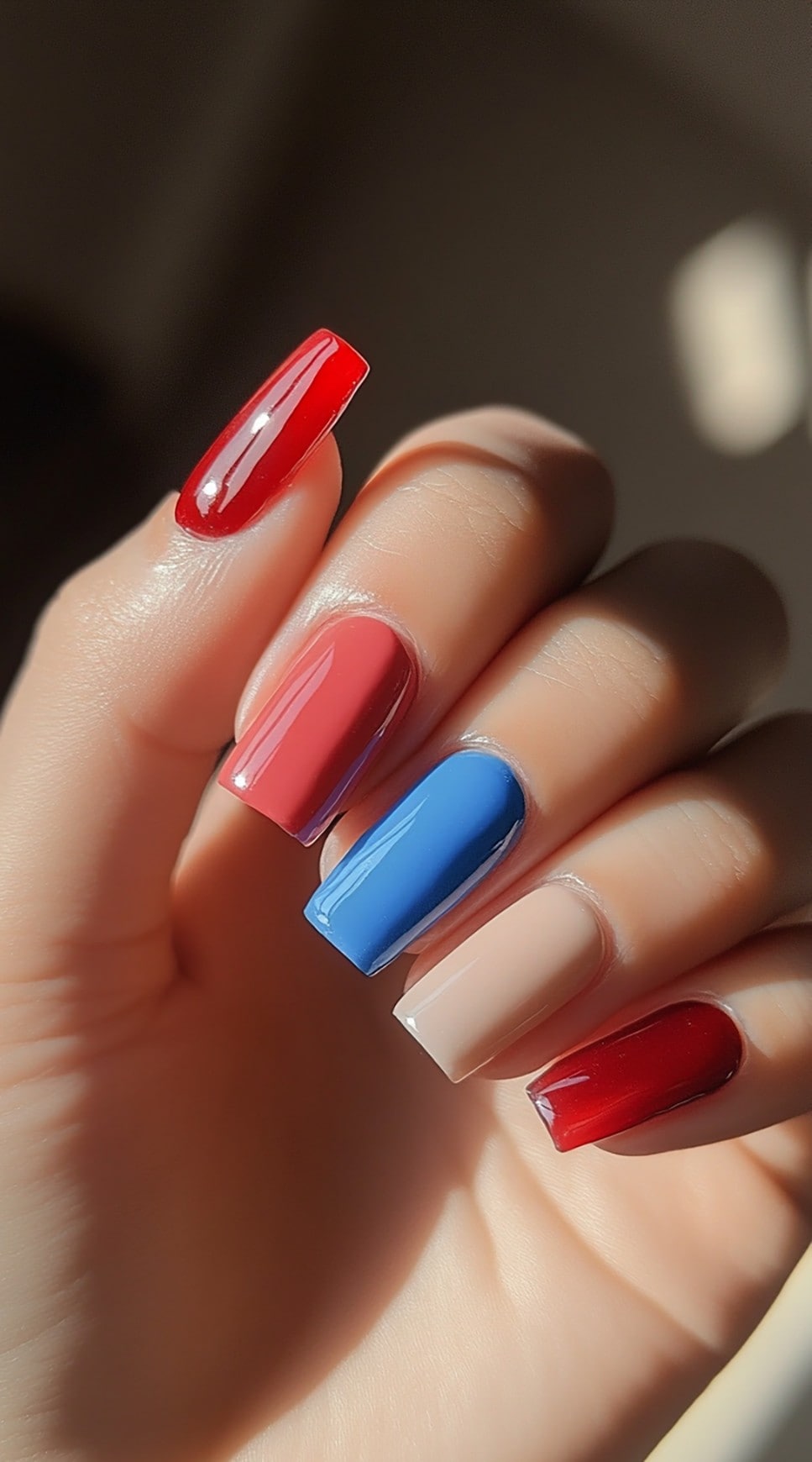 A hand with square-shaped nails painted in alternating glossy red, deep blue, and neutral tones for a simple yet stylish look.