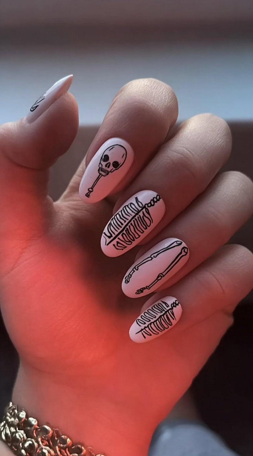 White almond nails with black hand-drawn outlines of various skeleton parts, including a skull, ribcage, and limbs.