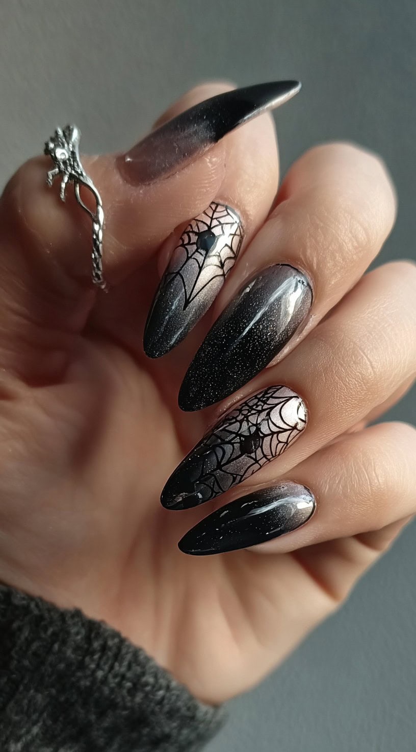 A hand with black-to-nude ombre nails, two of which feature spiderweb designs with black gemstone studs.