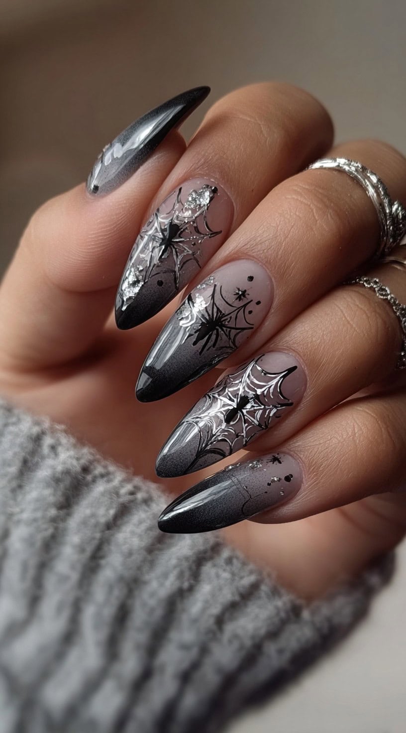 A hand with almond-shaped nails in silver-black ombre topped with black and metallic spiderwebs and glittering foil.