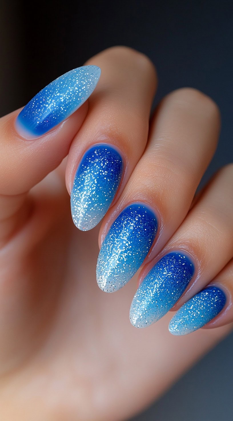 Almond-shaped nails with a vibrant blue to white ombre and soft silver glitter sprinkled across the entire surface.